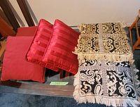 Seven pillows arranged on a glass table, showing four solid red pillows (two plain and two striped satin-like) and three patterned pillows with fringes.