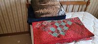 Quilt and blankets stacked on a bed with a visible mattress and wooden bed frame.