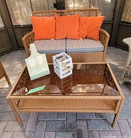 Rattan loveseat with orange cushions positioned behind the amber glass coffee table on a brick patio.
