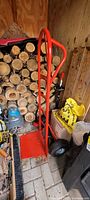 Full view of red metal dolly hand truck with large wheels standing against a woodpile indoors.