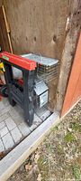 Two metal wire rodent cages stacked vertically near a wooden wall next to two red and black plastic saw horses.
