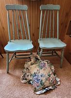 Two vintage green painted wooden chairs with spindle backs and turned legs, showing signs of wear. Two floral printed cushions placed in front.