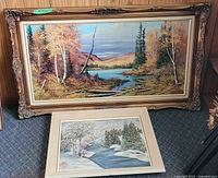 Photo showing both framed paintings: large ornate framed autumn landscape and smaller light-frame winter scene.