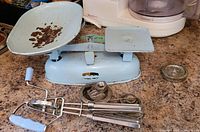 Full view of vintage blue kitchen scale with large rusty bowl and blue-handled manual hand mixer with chrome beaters placed in front.