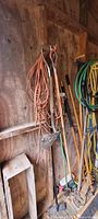 Wall with various garden tools hanging, includes shovels and hoes with wooden handles, coiled cables or extension cords