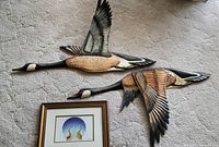 Two carved and painted wooden flying bird wall arts shown from above on carpet with framed abstract painting below.