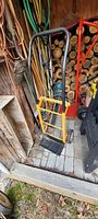 Yellow hand dolly standing upright next to various garden tools and firewood, showing the entire dolly.