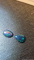 Two natural black opals on black textured background showing multicolored flashes of red, green, and blue colors under lighting.
