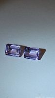 Pair of natural amethyst gemstones laid on a flat surface, showing rectangular emerald cut and pink-purple color.