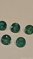 Close up of five round cut green emerald gemstones arranged on a beige surface, showing facets and some natural inclusions.