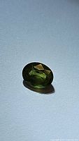 Close-up image of a natural green sapphire gemstone in oval cut on a white surface, displaying facets and clarity.