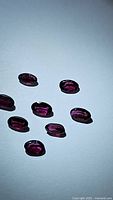 Seven oval cut deep purple-pink rhodolite garnet gemstones arranged on a white background showing clarity and faceting.