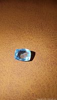 Close-up photo of a rectangular cushion-cut sky blue aquamarine gemstone placed on a brown textured surface, highlighting its color and inclusions.