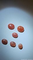 Six oval cabochon strawberry quartz gemstones arranged on a white surface under direct light, showing natural inclusions and pinkish hues.