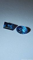 Photo showing two natural Swiss blue topaz gemstones, one rectangular cut and one oval cut, placed on a flat surface to display color and cut details.