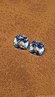 Pair of cushion cut baby blue topaz gemstones on brown textured surface, showing clarity and color.