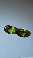 Two oval cut, vivid green natural peridot gemstones placed side by side on a light background showing clarity and color.