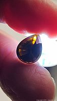 Photo of a pear-shaped deep dark orange-yellow tourmaline gemstone held between fingers with light transmitting through, showing facets and clarity.
