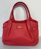 Front view of red pebbled leather tote bag with dual rolled handles and metal logo plate