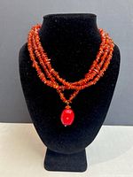 Three strands of amber beads arranged on a black mannequin neck, featuring a red geometric pendant as centerpiece.