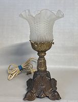 Photo of the lamp showing the frosted ruffled glass shade and ornate brass base with the cord coiled behind.