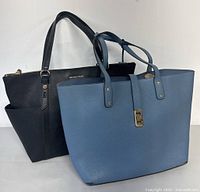 Pair of Michael Kors tote bags including a black zippered leather tote and a slate blue leather tote with gold turn-lock detail shown together