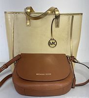 Front view showing the two Michael Kors bags, gold tote behind the brown crossbody, highlighting bag styles and sizes.