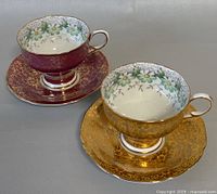 Pair of paragon teacups with matching saucers. One teacup/saucer set in deep burgundy with gold filigree; the other in gold with similar filigree. Both have floral interior design.