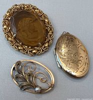 Three vintage gold-tone jewelry pieces: one large oval amber glass cameo brooch, one gold-tone engraved locket pendant, and one filigree oval brooch with pearl.