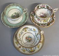 Overhead view of three porcelain teacup and saucer sets with different hand-painted floral and bird motifs, arranged on neutral background.