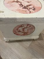 Shows front corner of rectangular wooden box. Side oval design depicts two embracing angels in a soft classical style with muted pink and brown tones. The box paint is distressed with some visible minor chips and wear.