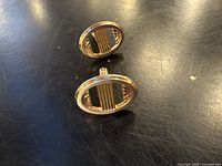 Two oval brass cuff links lying on a dark surface. The design includes vertical cut-out bars on circular fronts.