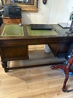 Full view of antique oak desk with green leather inset top, showing hand carved legs and wooden panels under desktop.