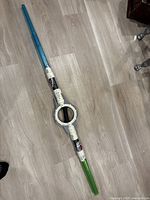 Full view of the dual-ended lightsaber placed on a wooden floor, showing blue and green translucent blades and detailed handle with circular center.