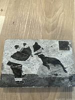 Front view of the rectangular soap stone sculpture showing an Inuit person reaching out to a seal in black inlay on a gray stone background, artist signature visible.