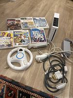 Nintendo Wii console bundle showing console upright, multiple Wii game cases, Wii Remote, Nunchuk, and Wii Wheel accessory on floor.