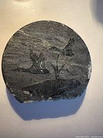 Front view of round, black and grey soapstone with carved geese flying over water and reeds.