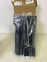 Two black flexible downspout extensions packaged in clear plastic bags, placed standing upright against a wall with a cardboard box behind.