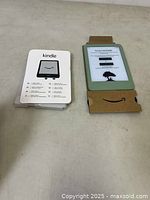 Front view of Kindle device with display on showing setup screen and accompanying cardboard packaging insert with Kindle instructions.
