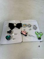 Photo of two cards with various Crocs charms attached, including a black bow, hearts, round silver charms, a pink car, clam shell with fish, and green frog face charm.