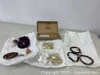 Assorted loose stones and minerals laid out on white paper, including polished brown stones, white spherical mineral stones, and rough mineral pieces in a brown cardboard box and a purple pouch.