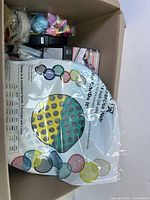 Top-view photo of a cardboard box filled with various small items including colorful pieces, tape rolls, and a packaged pet water dish.