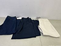 Three pairs of women's pants folded on a flat surface: dark blue denim jeans, black pants, and cream-colored jeans.