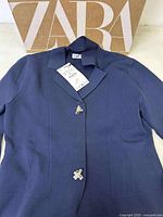 Navy blue Zara blazer jacket laid flat with original large Zara box behind it, showing front buttons and attached tag.