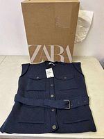 Navy blue Zara knit vest folded with original box in the background, showing buttons, pockets, belt, and tags