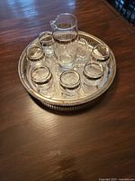 Glass cocktail pitcher with six matching glasses arranged on a silver finish round tray on a wooden table.