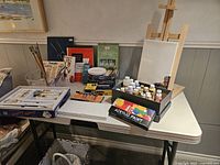 Full view of the table displaying multiple painting supplies including acrylic paints, brushes, sketch pad, easel with canvas
