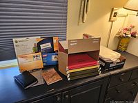 Office supplies displayed on black dresser including colored hanging file folders in box, label packages, and planner book.