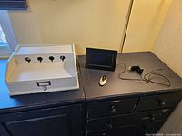 White Pottery Barn charging station with four charging ports and storage drawer on the left, 7-inch digital photo frame with remote and power adapter on the right on top of a black dresser.