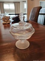 Front angle view showing the glass compote with lid featuring the Three Graces figure knob, placed on a wooden table.